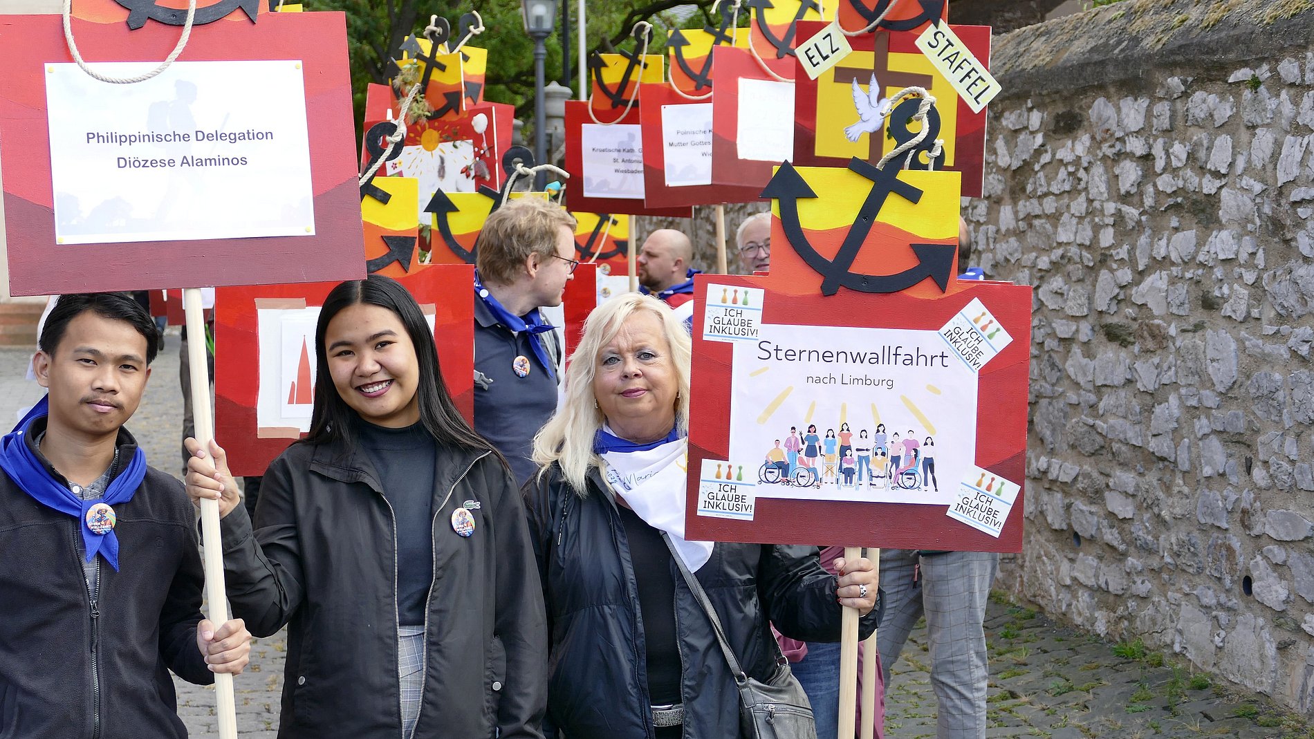 Eine Gruppe von Personen, vermutlich Teilnehmer an einer Wallfahrt, die verschiedene Plakate hochhalten. Die Plakate haben Text und Abbildungen. Einige der Plakate sind mit Namen wie "Philippinische Delegation", "Diozee Alaminos", "Sternenwallfahrt nach Limburg" und "IHQ GLAUBE INKLUSIV" versehen. Es gibt auch Abbildungen von Menschen, von denen einige in traditioneller Kleidung zu sein scheinen. Der Hintergrund zeigt eine Steinmauer und einen Teil eines Gebäudes.